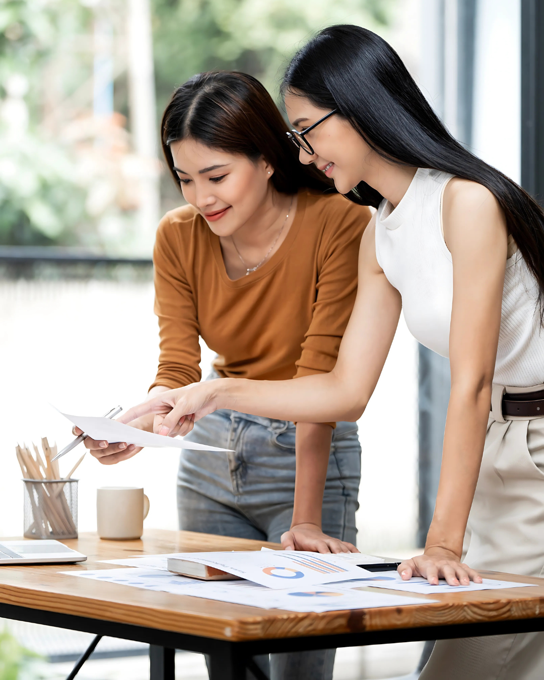 Happy-Asian-Businesswomen-Collaborating-on-Outsourced-Financial-Services (1) Two happy Asian businesswomen collaboratively reviewing documents and charts, demonstrating efficient coworking for outsourced financial services.